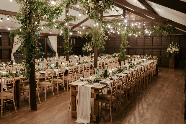 The inside of a barn decorated with long tables, greenery and fairylights