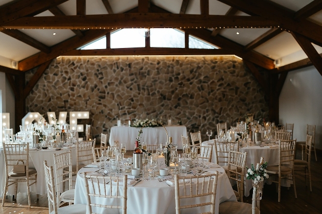 The inside of a room with wooden beams, windows, a stone wall and round tables decorated with flowers