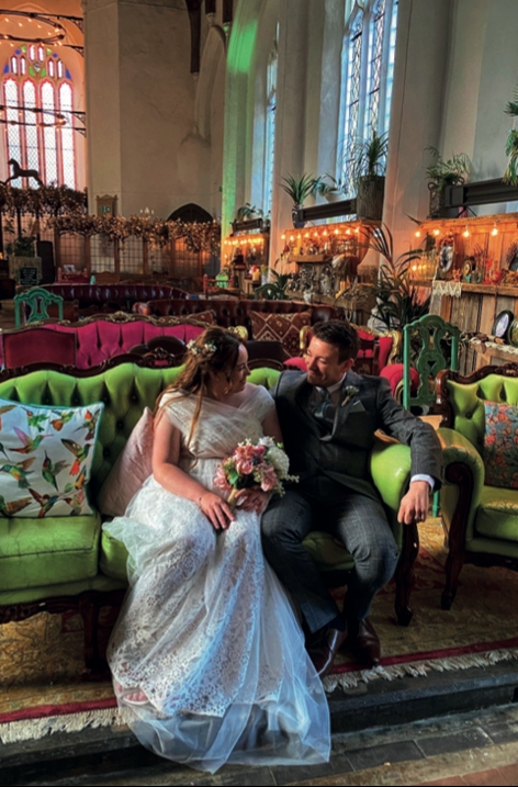 A bride and groom sitting on a colourful sofa surrounded by plants and fairylights