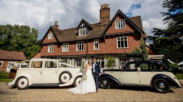 A bride and groom standing outside of a brick building with two white cars